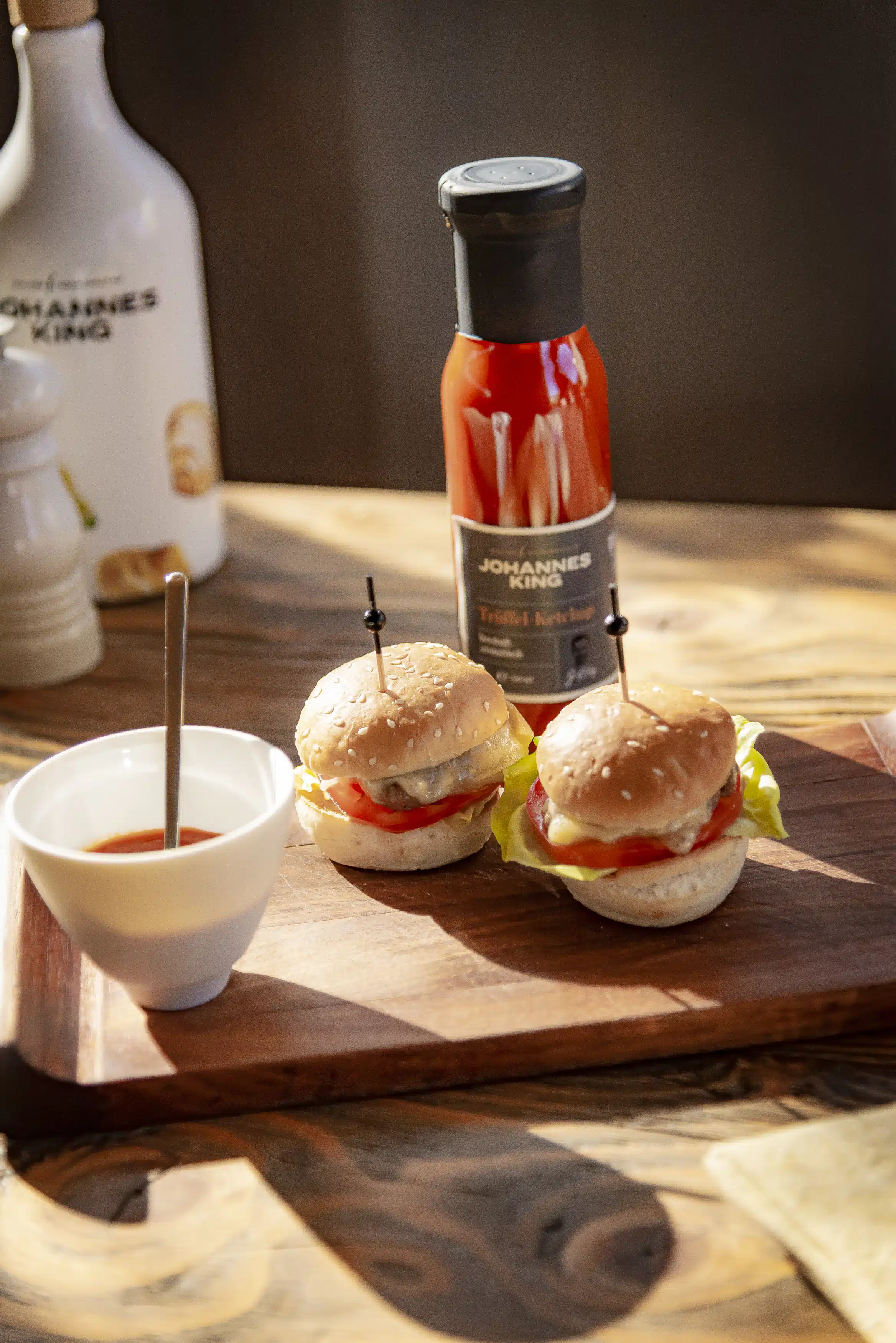 Two small hamburgers with lettuce and tomato sit on a wooden board next to a bowl of sauce. A bottle of Johannes King ketchup, perfect for any content marketing agency's food shoot, stands in the background on a rustic wooden table.