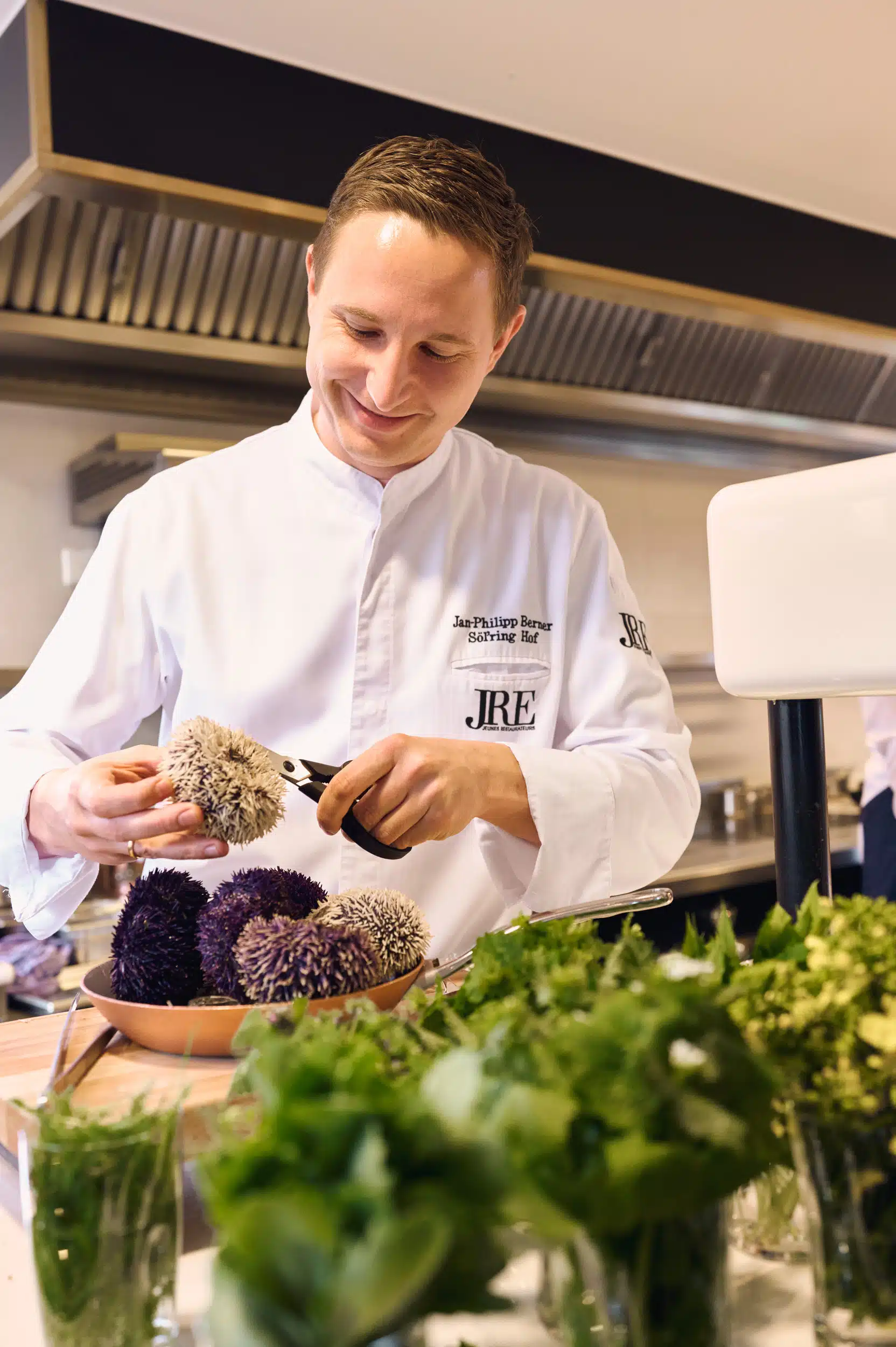 Ein Koch in weißer Uniform lächelt, während er in einer professionellen Küche Seeigel zubereitet. Im Vordergrund sind frische Kräuter zu sehen, im Hintergrund Metallregale.