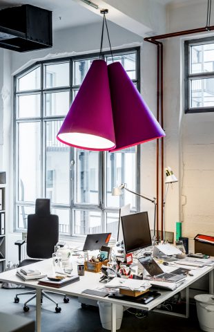 Ein moderner Büroraum mit großen Fenstern, überfüllten Schreibtischen mit Computern und Büromaterial, Bürostühlen und zwei übergroßen violetten Pendelleuchten – perfekt für ein Brainstorming zum Marketingkonzept eines Restaurants.