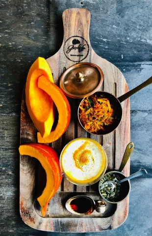 Auf einem hölzernen Servierbrett liegen rohe Kürbisscheiben, ein Topf mit geraspeltem Gemüse, eine Schüssel Suppe mit Schaum, ein Schälchen mit Samen und ein kleiner Behälter mit dunkler Flüssigkeit – eine elegante Präsentation, perfekt für eine Hotelberatung. Der Hintergrund ist eine dunkle, strukturierte Oberfläche.
