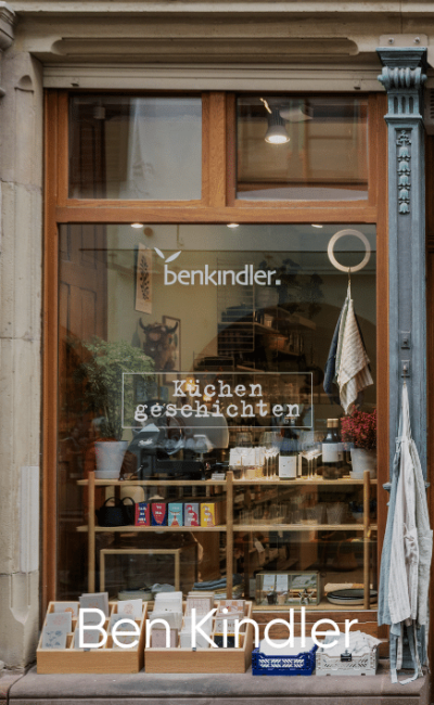 Im Schaufenster von Ben Kindler werden Bücher, Küchenartikel und Haushaltswaren ausgestellt. Die Worte „Küchengeschichten“ und „benkindler“ sind auf dem Glas zu sehen. Durch das Fenster sind Regale und Pflanzen zu sehen.