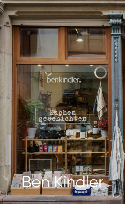 Im Schaufenster von Ben Kindler werden Bücher, Küchenartikel und Haushaltswaren ausgestellt. Die Worte „Küchengeschichten“ und „benkindler“ sind auf dem Glas zu sehen. Durch das Fenster sind Regale und Pflanzen zu sehen.