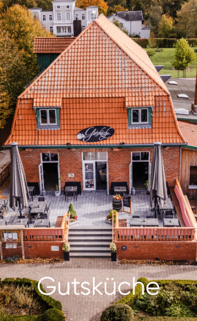 Luftaufnahme eines Backsteinrestaurants mit rotem Ziegeldach, Sitzplätzen im Freien, großen Sonnenschirmen und einem Schild mit der Aufschrift „Gutshof“ über dem Eingang. Im Hintergrund sind Bäume und Gebäude zu sehen. Unten steht das Wort „Gutsküche“.