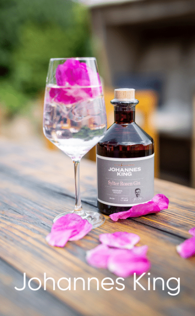 Ein Glas mit einem klaren Getränk und rosa Rosenblättern steht neben einer braunen Flasche mit der Aufschrift „Johannes King Sylter Rosen Gin“ auf einem Holztisch mit verstreuten Rosenblättern. Unten ist der Text „Johannes King“ zu lesen.