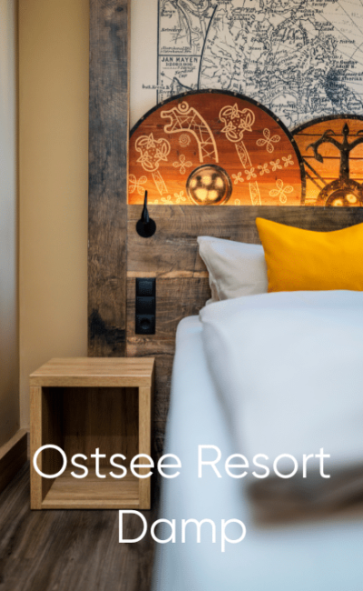 Eine Nahaufnahme eines gemachten Bettes mit weißen und gelben Kissen, einem hölzernen Kopfteil mit einem Kartendesign, einem kleinen hölzernen Nachttisch und dem darüber gelegten Text „Ostsee Resort Damp“.