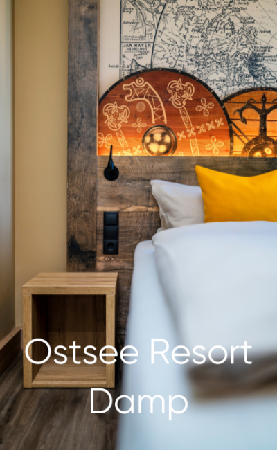 Eine Nahaufnahme eines gemachten Bettes mit weißen und gelben Kissen, einem hölzernen Kopfteil mit einem Kartendesign, einem kleinen hölzernen Nachttisch und dem darüber gelegten Text „Ostsee Resort Damp“.