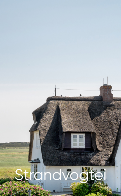 Ein weißes Häuschen mit Strohdach und Dachgaube steht vor offenen grünen Feldern unter klarem Himmel. Am unteren Bildrand ist das Wort „Strandvogtei“ zu sehen.