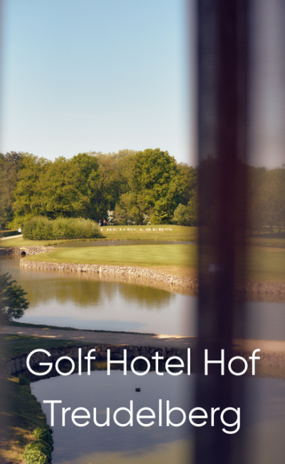 Blick durch ein Fenster auf einen Golfplatz mit einem Teich und grünen Bäumen im Hintergrund. Unten steht der Text: „Golfhotel Hof Treudelberg“.