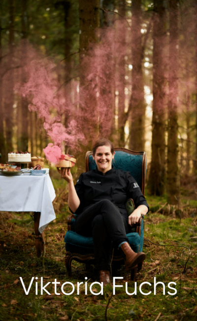 Eine Frau in einer schwarzen Kochjacke sitzt auf einem blauen Stuhl im Wald und hält eine Torte in der Hand, aus der rosa Rauch aufsteigt. Neben ihr steht ein Tisch mit Kuchen und Gebäck. Unten steht der Text „Viktoria Fuchs“.