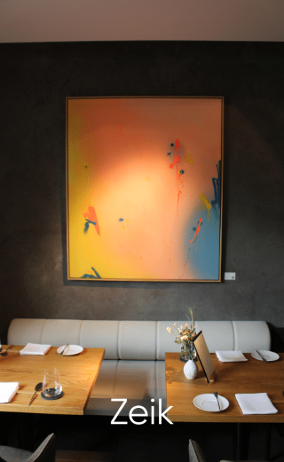 Ein modernes Restaurant mit einem Holztisch für vier Personen, einer weißen Sitzbank und einem großen abstrakten Gemälde in Orange-, Gelb- und Blautönen an einer dunklen Wand. Unten ist der Text „Zeik“ zu sehen.