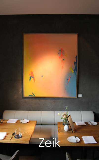 Ein modernes Restaurant mit einem Holztisch für vier Personen, einer weißen Sitzbank und einem großen abstrakten Gemälde in Orange-, Gelb- und Blautönen an einer dunklen Wand. Unten ist der Text „Zeik“ zu sehen.