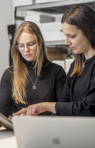 Two women stand at a desk in an office, one holding a tablet and the other pointing at a laptop screen. Shelves with boxes and jars are visible in the background.