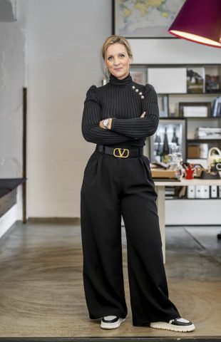 A woman stands with arms crossed in a modern office. She wears a black sweater, wide black pants, and white-black shoes. Behind her are desks, shelves with books, and large purple pendant lights.