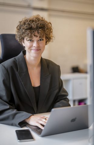 A person with curly hair sits at a desk working on online shop programming on their laptop. They are wearing a dark blazer and smiling at the camera. A smartphone is placed on the desk next to the laptop.
