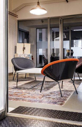 Ein moderner Büro-Loungebereich mit drei Stühlen um einen weißen Couchtisch, einem gemusterten Teppich, Glaswänden und einer Deckenleuchte – perfekt für informelle Meetings oder Marketing-Schulungen. Im Hintergrund ist ein separater Arbeitsbereich mit einem roten Tisch zu sehen.