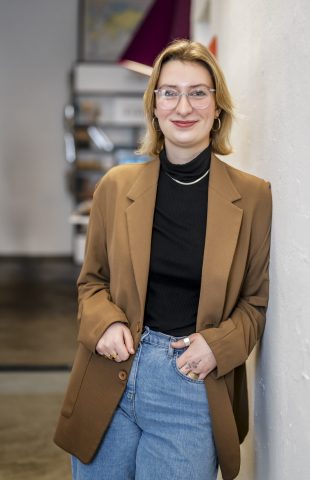 A person with short blond hair wearing glasses, a brown blazer, black turtleneck, and blue jeans stands indoors, leaning against a white wall and smiling at the camera. Shelves filled with seo texts are visible in the background.