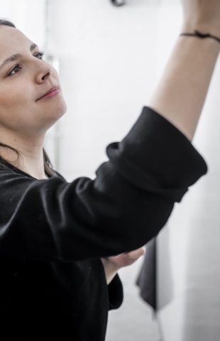A woman with long brown hair wearing a black top is writing or drawing on a whiteboard in a bright office space. She appears focused on her task.