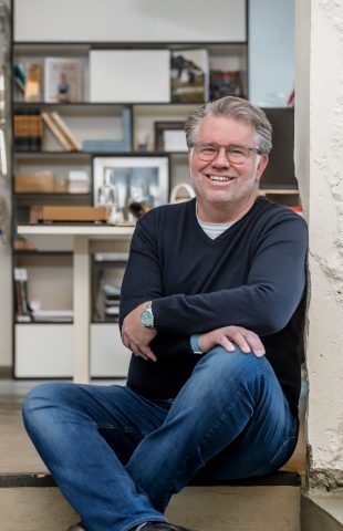 A man with gray hair and glasses, wearing a navy sweater and blue jeans, sits on the floor indoors, smiling, with his arm resting on his knee. Behind him are shelves with books and framed photos, including one of vita antonia wien and lars ammer.