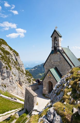 A small stone chapel with a green roof sits on a mountain slope, surrounded by rocky hills and greenery under a bright blue sky. A few people, perhaps exploring for tourism, are near the entrance, and a winding path leads up to the chapel.