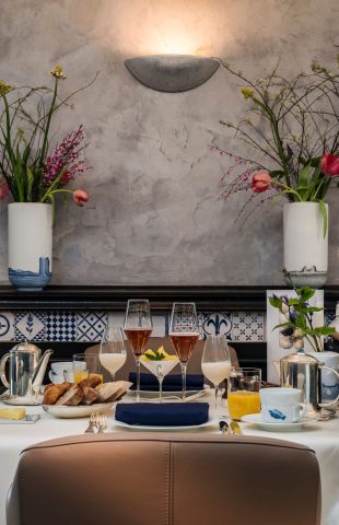 A table set for a meal with various dishes, pastries, drinks, and glassware—perfect for restaurant marketing—sits between two white vases with flowers against a textured wall, with a display case holding cured meats to the right.