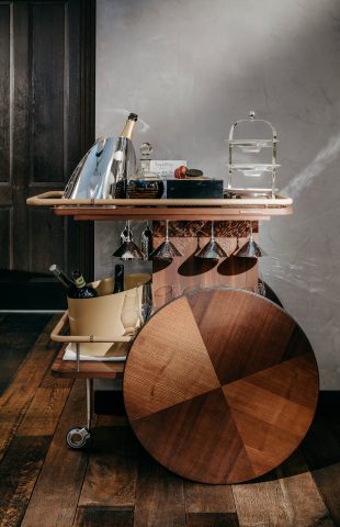 A wooden bar cart, perfect for restaurant marketing, features a segmented round tray, wine bottles in a bucket, an ice bucket, glasses below the counter, stacked pastry trays, and drink accessories—all set on a hardwood floor.